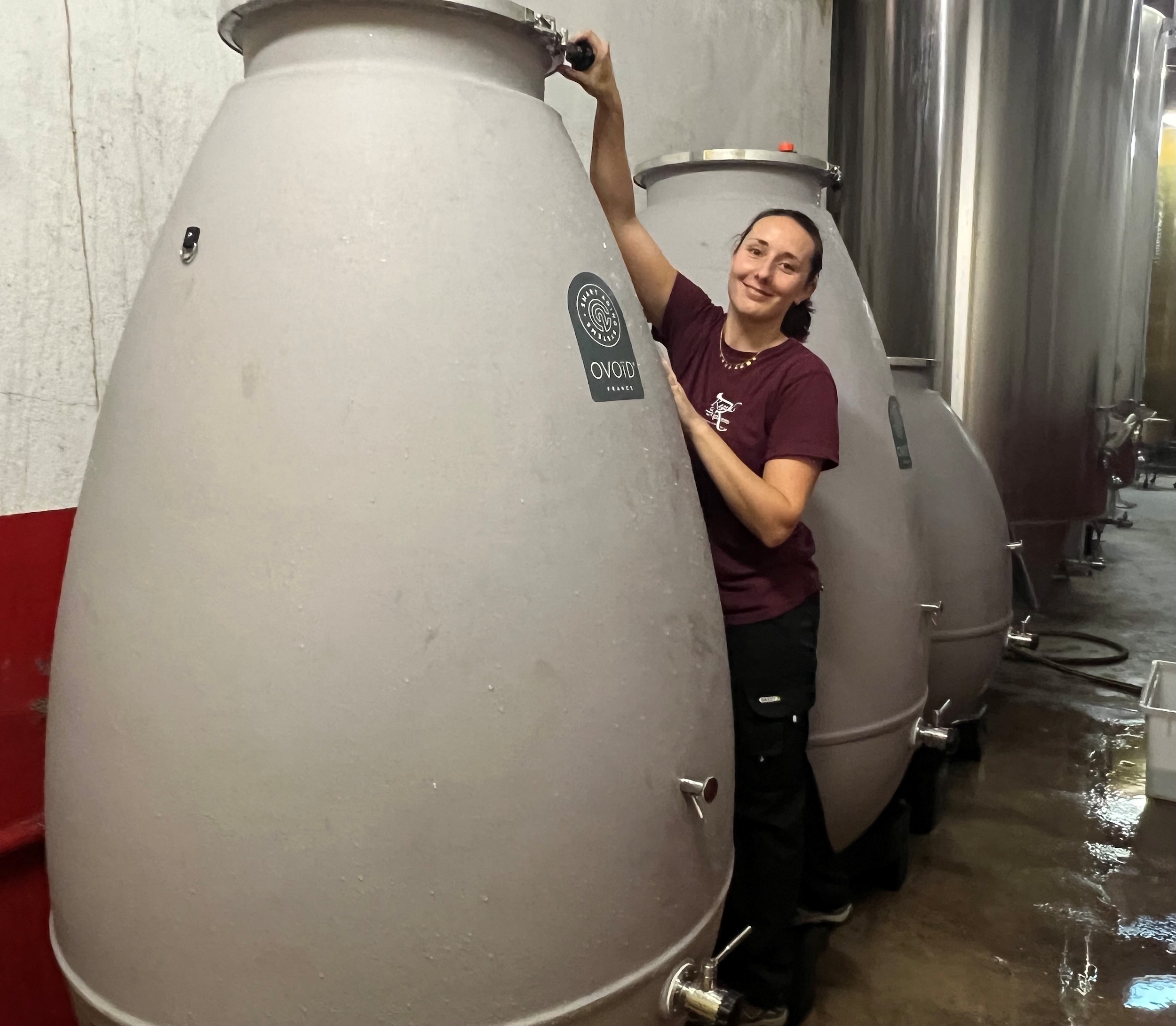 Winemaker smiling between concrete egg-shaped fermentation tanks in a wine cellar.