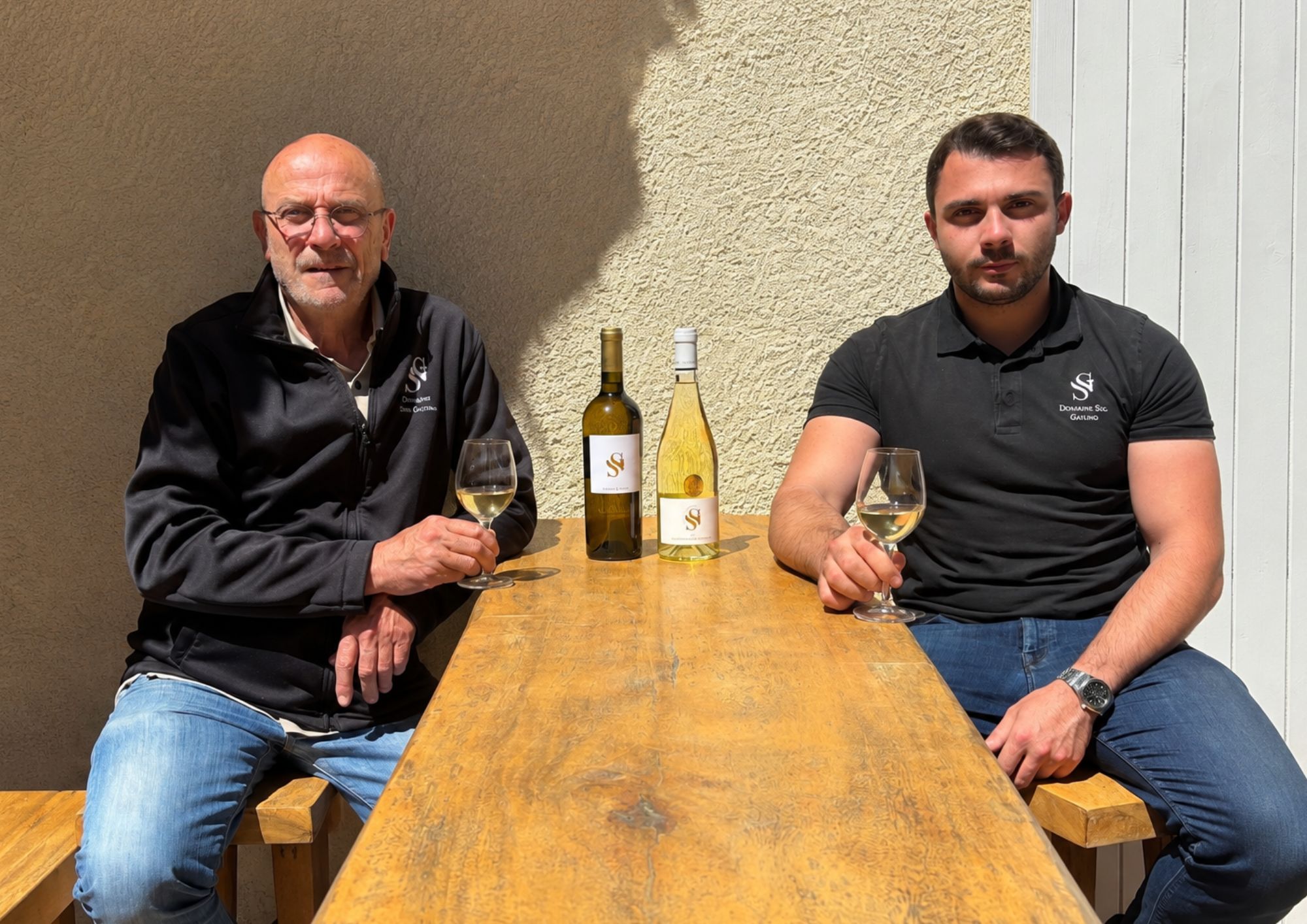 Two winemakers from Domaine Sec Gailino sitting at a table with glasses of white wine and two bottles bearing their SG label.