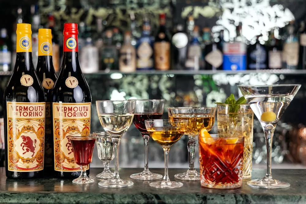 Bottles of Vermouth di Torino next to vermouth cocktails and glasses set out on a bar counter