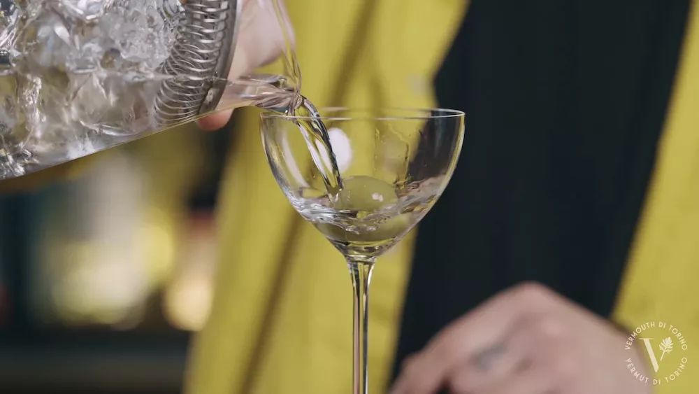 Clear cocktail being poured from a mixing glass into a stemmed coupe, close-up against a yellow and black background