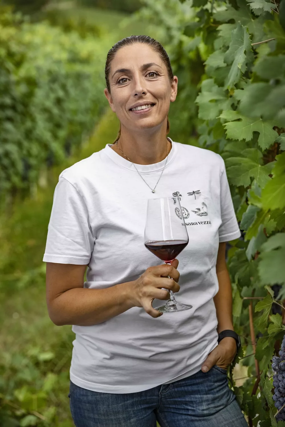 Sara Vezza of the Saffirio estate, smiling in a white t-shirt with a glass of red wine among the vines