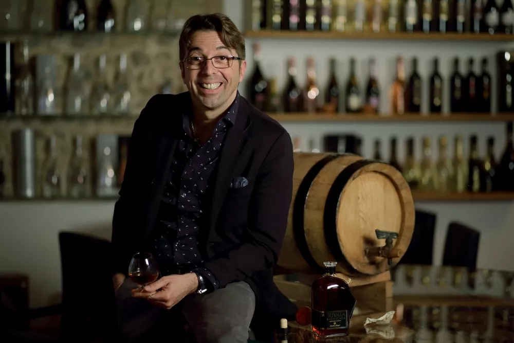 Stefano Bottega smiling with a glass in hand, seated by a small barrel in front of shelves of bottles
