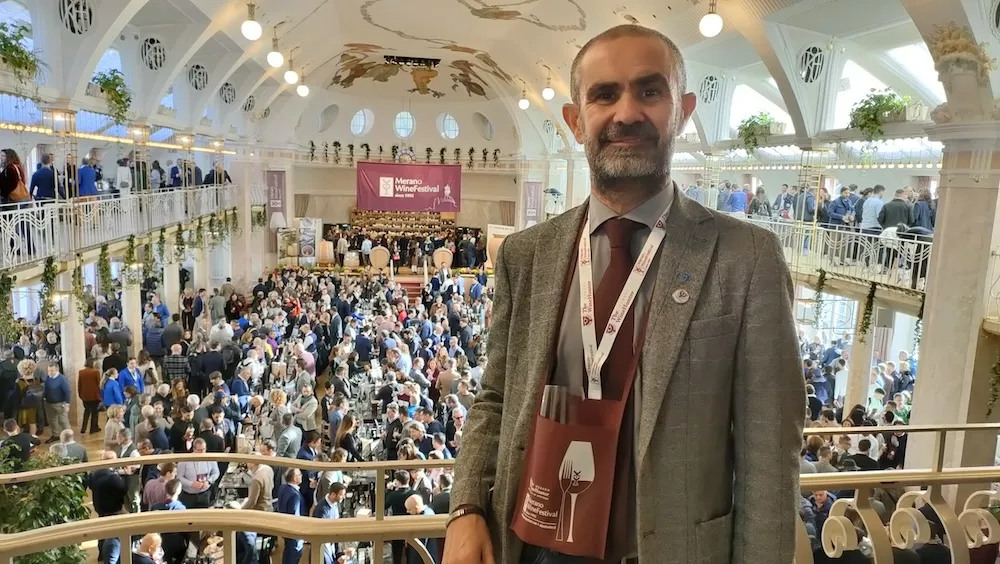 Bearded man in a grey jacket with a lanyard in front of the packed hall of a large wine fair